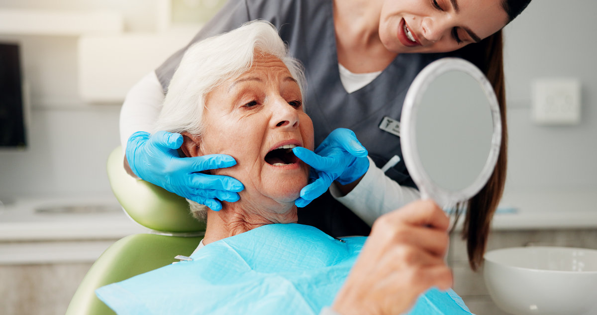 Dentist, senior woman and consulting with mirror for teeth cleaning, dental implant or oral hygiene. Inspection, healthcare or doctor with old patient in clinic for mouth exam or root canal treatment.