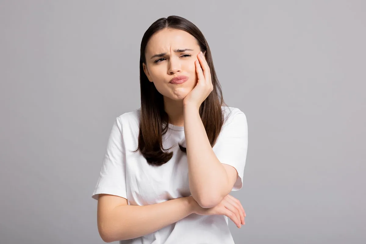 Crying tired sad woman suffers from painful toothache dental illness pointing on teeth standing on grey background in studio isolated wearing basic white t-shirt.