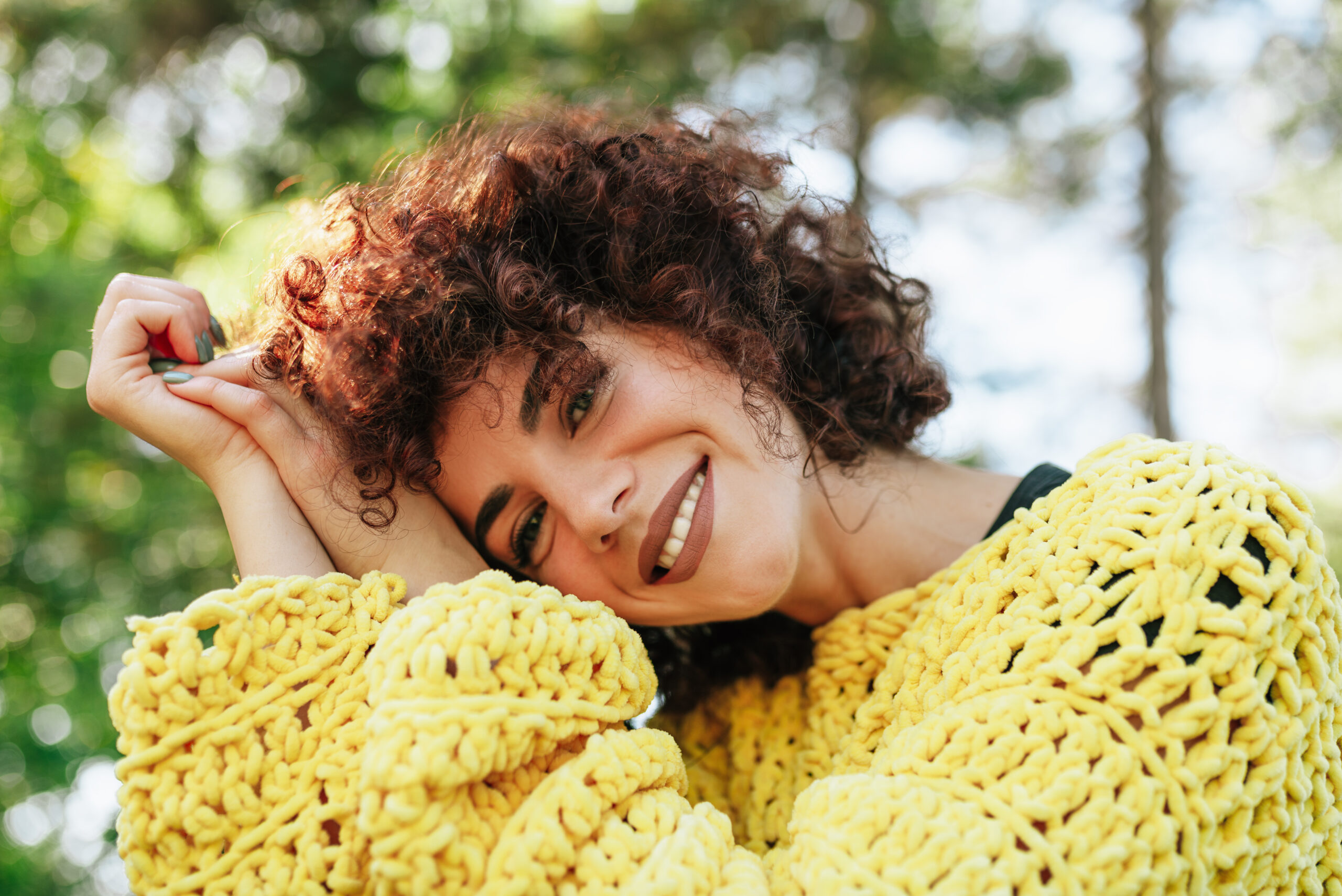 Close-up outdoor portrait of attractive young woman with curly hair and smiling. Female has positive expression, wearing knitted yellow sweater and posing against nature background. People, lifestyle