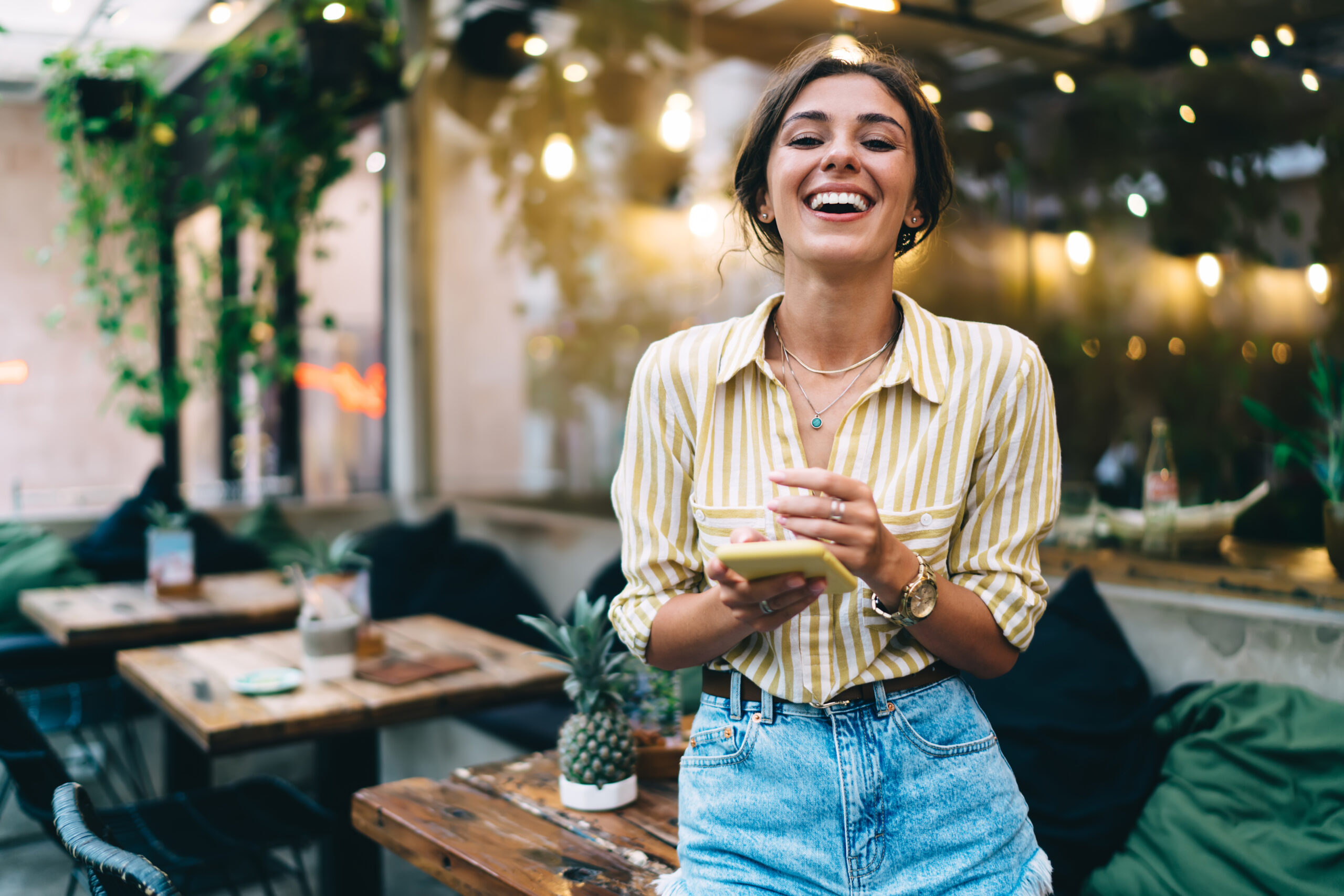 Smiling female employee with pendant on neck checking email on cellphone while working online in street cafeteria and looking at camera