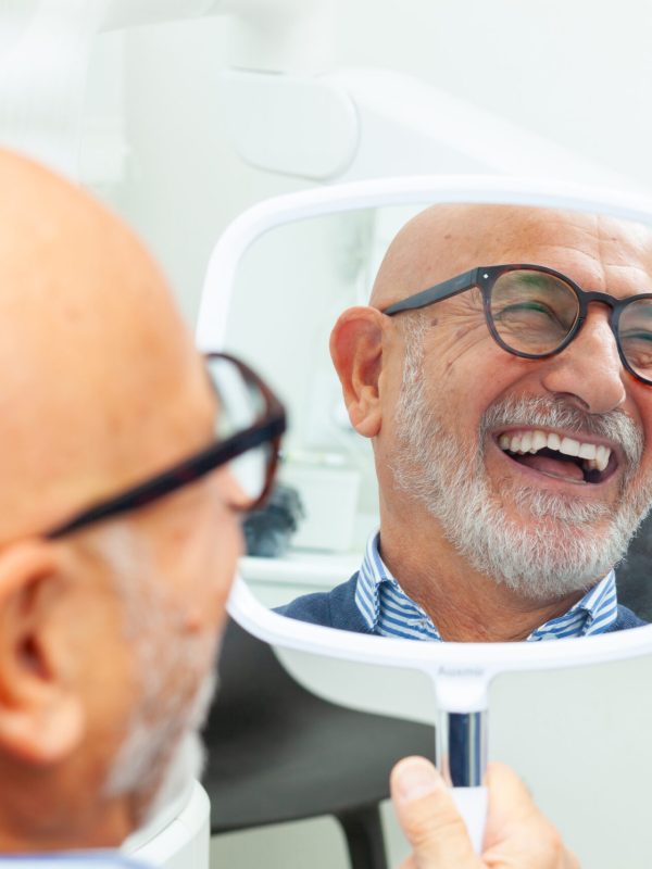 Happy senior patient admiring his perfect smile after a dental implant treatment, holding a mirror in his hand at the dental clinic