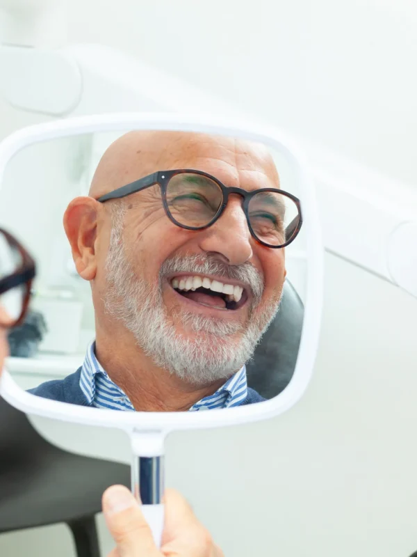 Happy senior patient admiring his perfect smile after a dental implant treatment, holding a mirror in his hand at the dental clinic