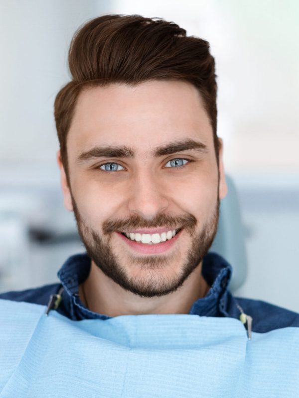 Cheerful bearde man patient sitting in dental chair and smiling at camera, copy space. Positive millennial guy showing his beautiful white smile after treatment at modern dental clinic