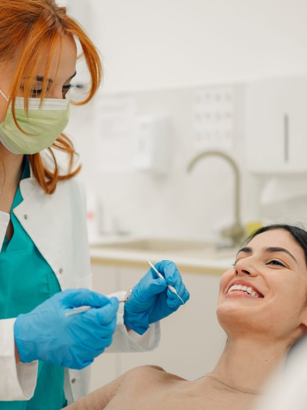 Female dentist wearing mask and gloves performing dental checkup cleaning teeth of happy woman patient in modern clinic, dental health concept