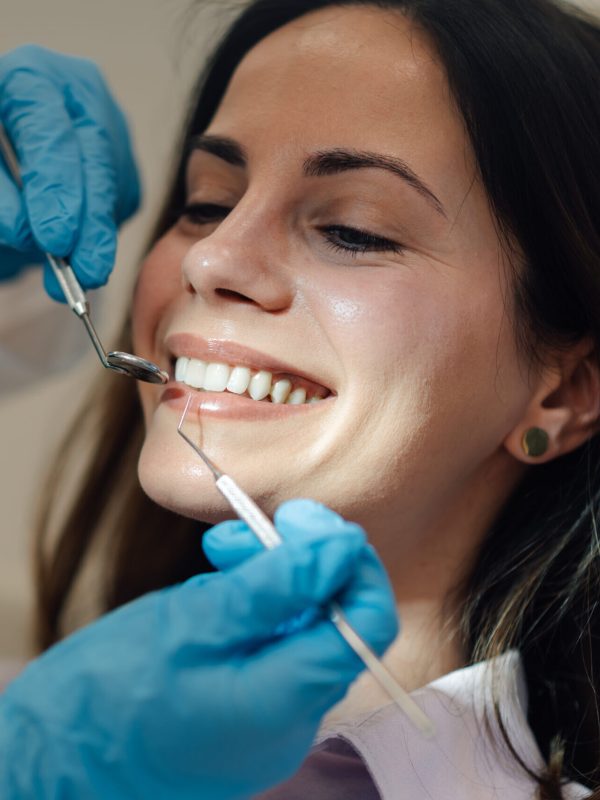 Dentist wearing blue gloves examining teeth of smiling young woman during a thorough dental checkup in a bright, clean clinic, ensuring optimal oral health and hygiene