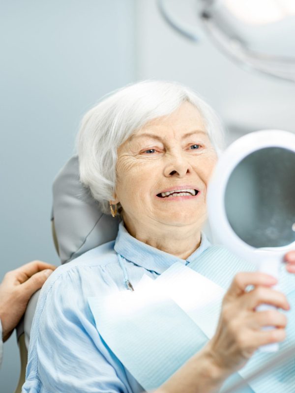 Happy elderly woman enjoying her beautiful toothy smile looking to the mirror in the dental office