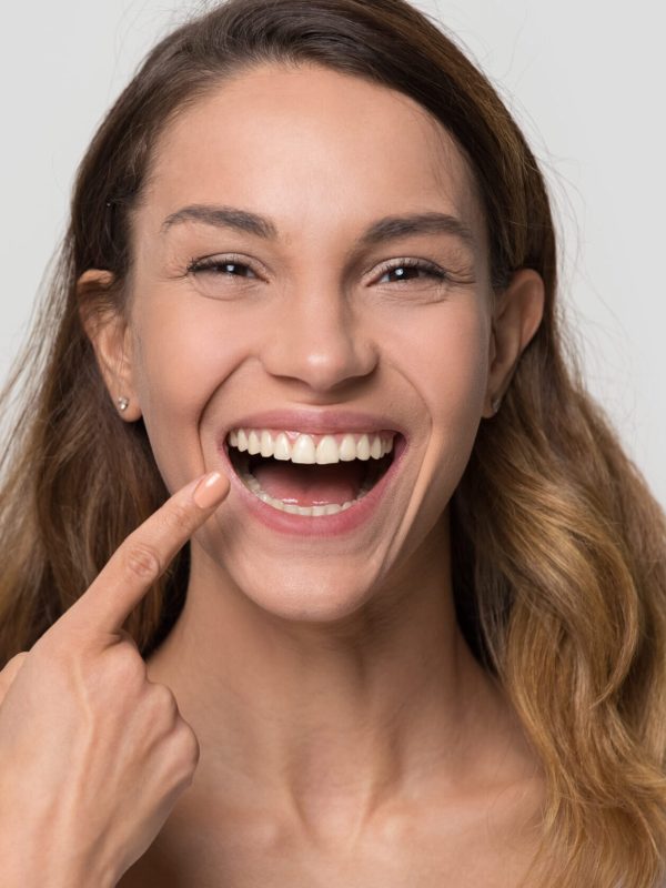 Happy young woman with white straight teeth perfect dent orthodontic smile pointing at tooth looking at camera isolated on studio blank background, dental health stomatology service concept, portrait