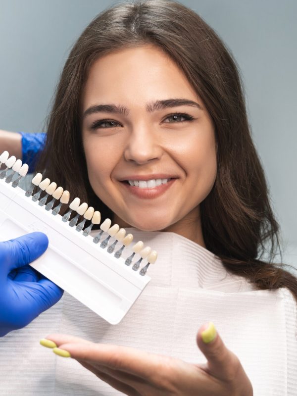 smiling brunette woman patient having appiontment in dental clinic picking up shade using tooth enamel scale held by dentist in blue gloves