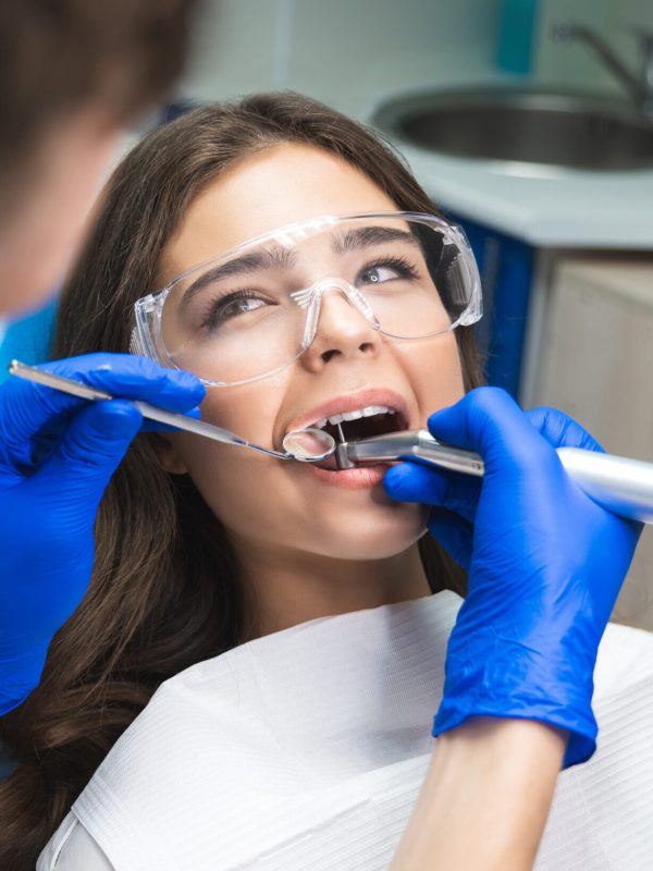 dentist in mask filling the patient's root canal while she is lying on dental chair in safety glasses under the medical lamp in clinic.