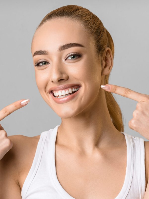 Orthodontic concept. Happy girl showing her beaming white teeth with two forefingers, grey background