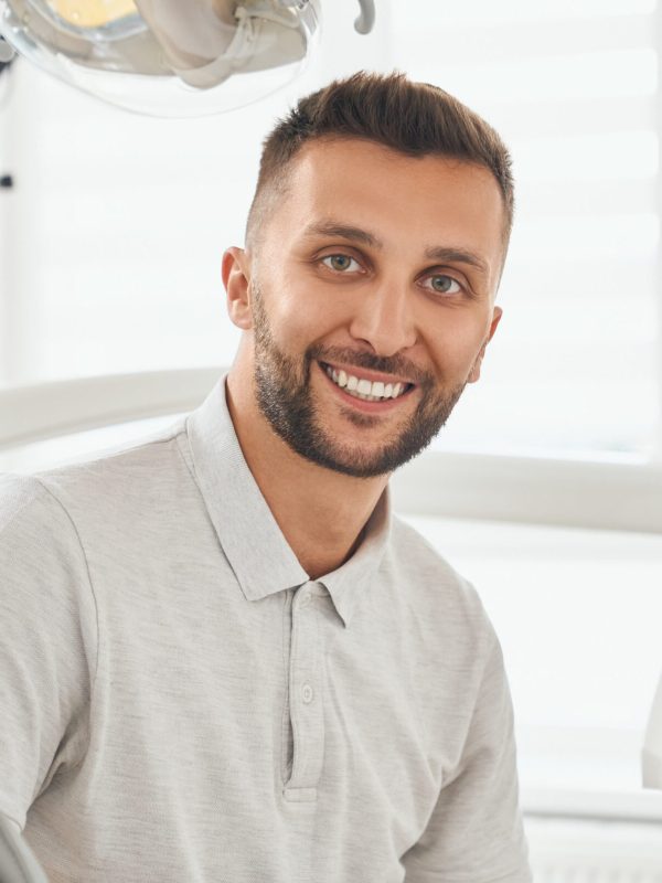 Portrait of happy bearded man in grey t-shirt sitting in dental chair, smiling and looking at camera. Young male patient looking satisfied after hygiene procedure at dental clinic