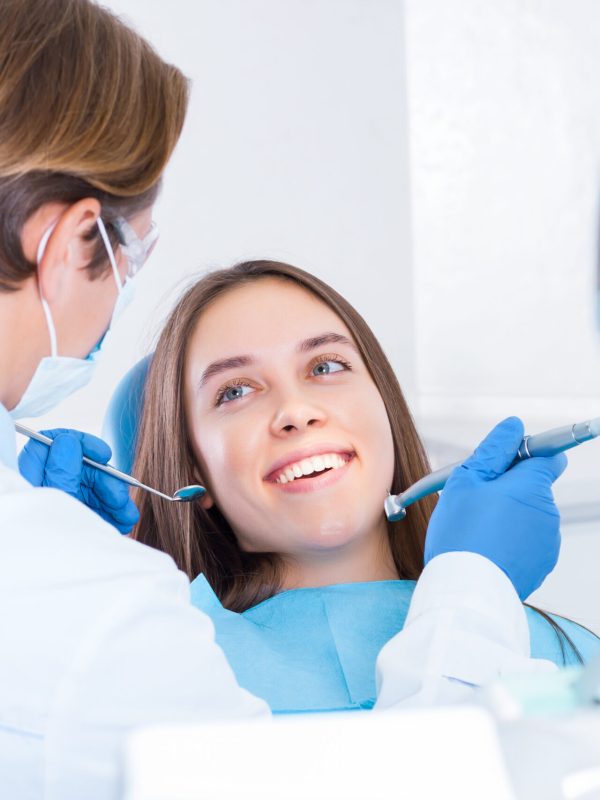 Smiling young woman is sitting in dental blue chair in clinic, office. Man doctor, orthodontist is conducting examination, treating teeth with tools, instruments. Visit to dentist concept.