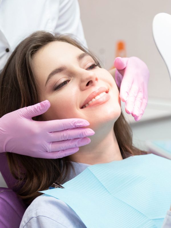 Young glad smiling woman patient looking at mirror after teeth whitening procedure, caries treatment, toothache elimination. Pretty happy girl at stomatologist appointment in dental clinic