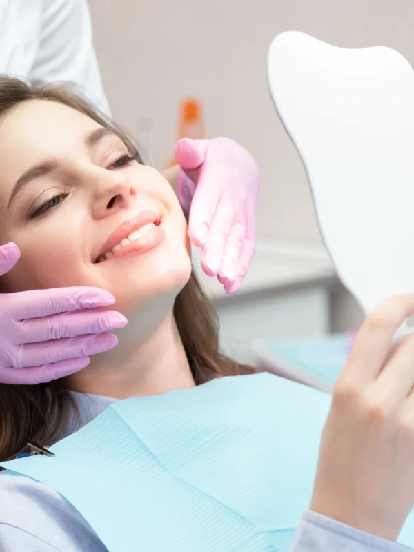Young glad smiling woman patient looking at mirror after teeth whitening procedure, caries treatment, toothache elimination. Pretty happy girl at stomatologist appointment in dental clinic