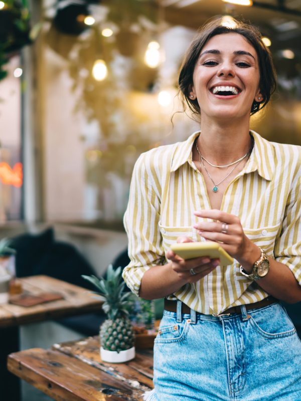 Smiling female employee with pendant on neck checking email on cellphone while working online in street cafeteria and looking at camera