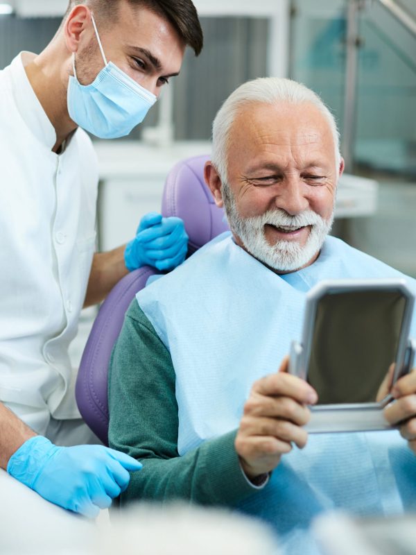 Satisfied senior man looking his teeth in a mirror after dental procedure at dentist's office.