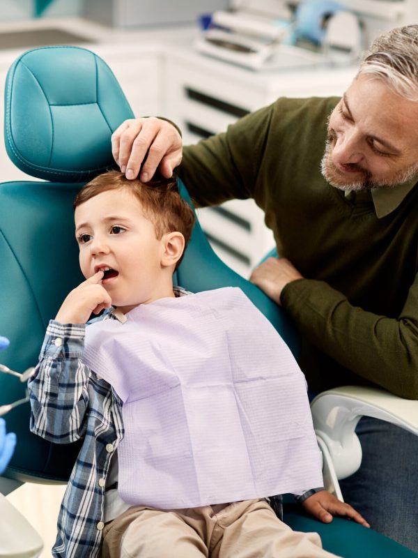 Little boy complaining of toothache to his dentist while being with his father at dental clinic.