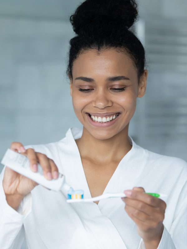 Happy young African American woman squeezing mint toothpaste from tube on plastic toothbrush for brushing teeth in bathroom, keeping morning routine for dental care, healthy enamel, mouth hygiene
