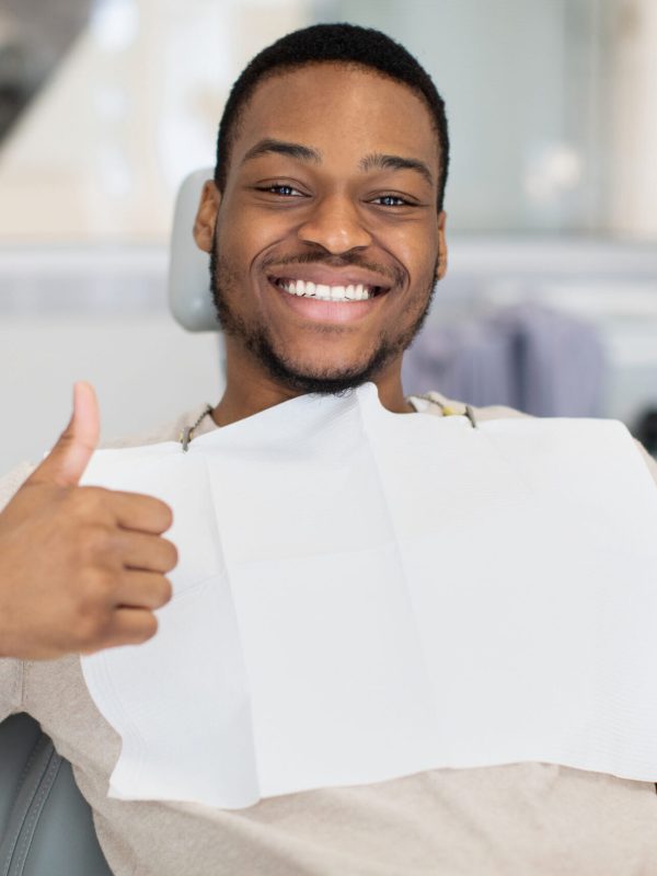 Happy young black man sitting in dentist chair, showing thumb up and smiling at camera. Handsome african american male patient enjoying quality dental services in modern stomatological clinic