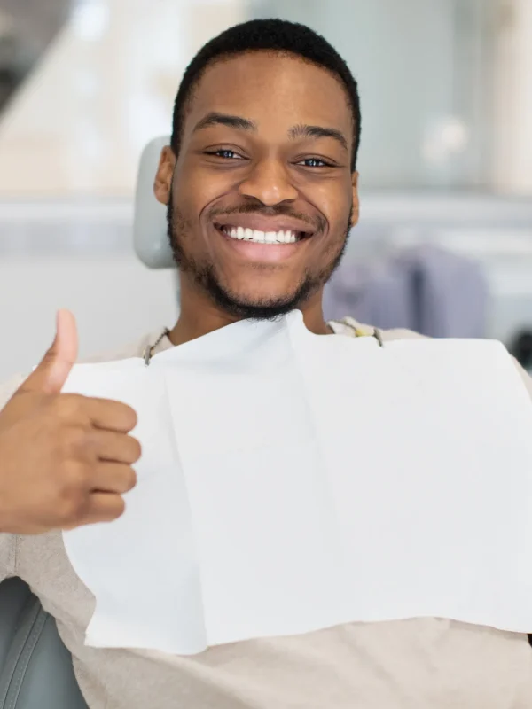 Happy young black man sitting in dentist chair, showing thumb up and smiling at camera. Handsome african american male patient enjoying quality dental services in modern stomatological clinic