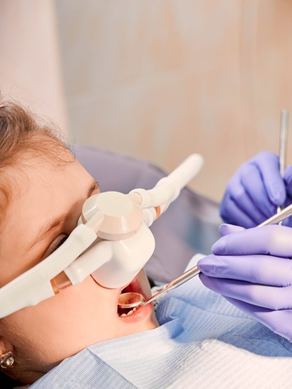 Female dentist checking child teeth with dental explorer and mirror while girl lying in dental chair with inhalation sedation at dental office. Concept of pediatric, sedation dentistry.