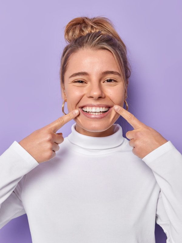 Dental care concept. Happy young fair haired woman points index fingers at toothy smile shows wel cared white teeth after whitening or dentist visit dressed casually isolated over purple background