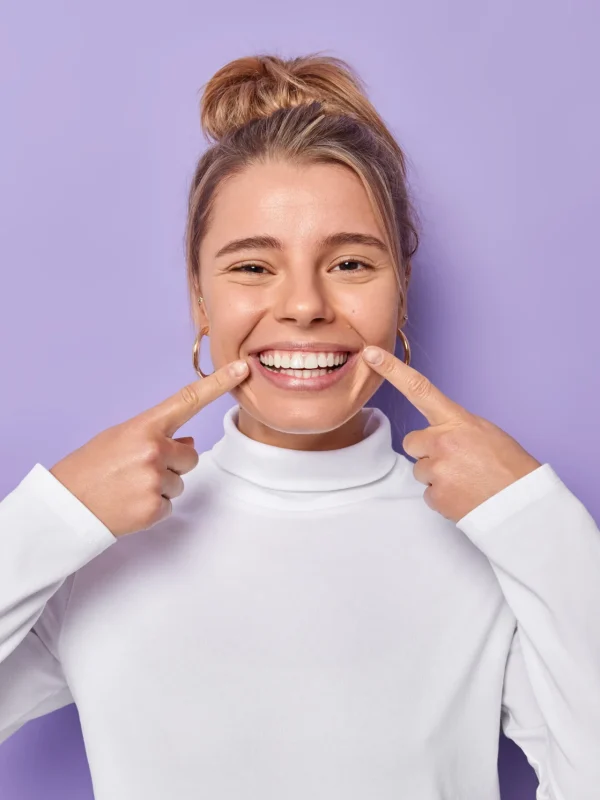 Dental care concept. Happy young fair haired woman points index fingers at toothy smile shows wel cared white teeth after whitening or dentist visit dressed casually isolated over purple background
