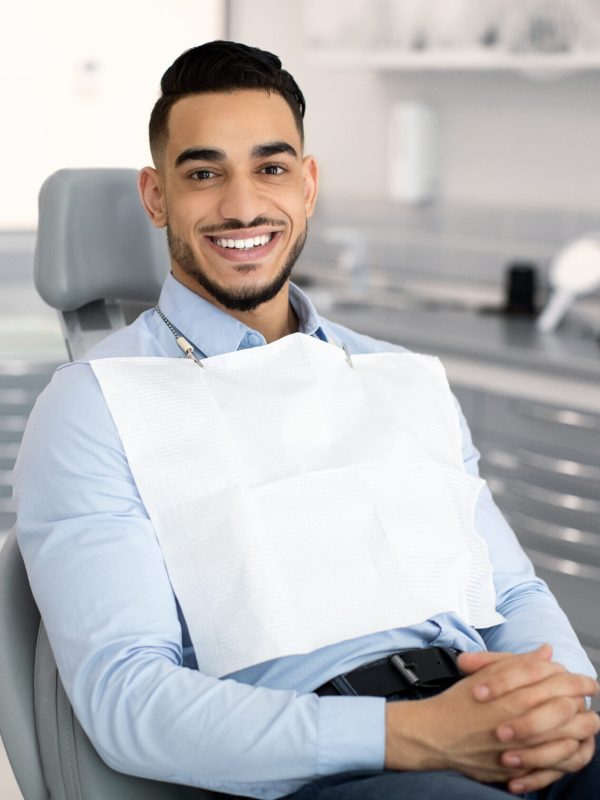 Stomatological Clinic. Portrait Of Handsome Arab Male Patient Sitting In Dentist Chair, Waiting For Check Up In Modern Cabinet, Young Middle Eastern Man Looking And Smiling At Camera, Copy Space