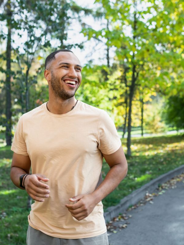 Cheerful and successful hispanic man jogging in the park, man running on a sunny day, smiling and happy having an outdoor activity.